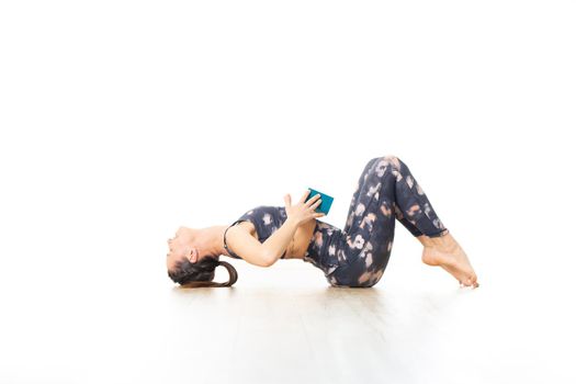 Young sporty attractive woman in bright white yoga studio, stretching and relaxing during restorative yoga using gadget. Healthy active lifestyle.