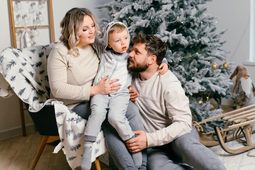 Christmas Family Happiness Portrait of dad, pregnant mom and little son sitting armchair at home near Christmas tree hug smile European young adult family holiday morning