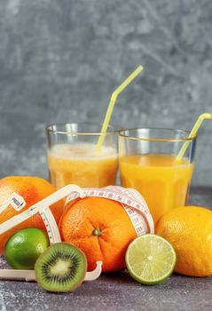 An orange wrapped with a measuring tape and a caliper surrounded by fresh fruits and glasses of juice and smoothies on a gray concrete background. The concept of slimming, bring the figure into shape.