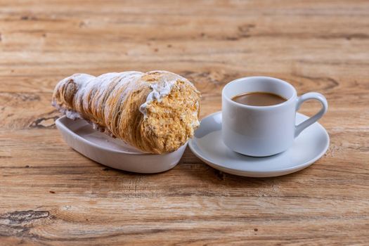 A cup of coffee next to a delicious aromatic puff pastry with protein cream, a delicious breakfast or snack on wooden table.