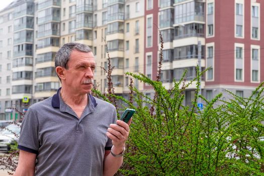 Adult caucasian senior man in a T-shirt stands against the background of residential buildings in the city in spring or summer and holds a smartphone. Selective focus.