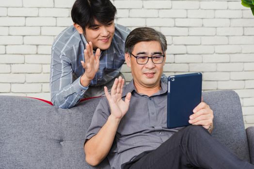 Asian senior businessman with digital tablet discuss together with young team in office. Father man and his son sit on sofa talking chatting on video call conference on tablet in living room at home