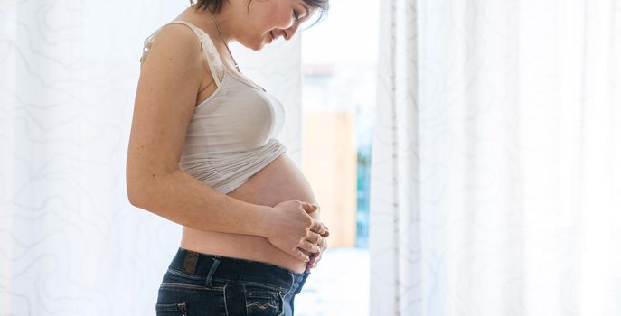 Happy Caucasian pregnant mother touching her tummy, blue jeans, close up