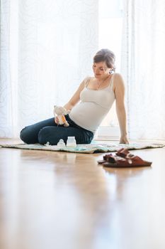 Happy Caucasian pregnant mother is sitting on the floor, touching her tummy, blue jeans