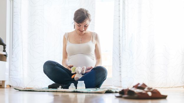 Happy Caucasian pregnant mother is sitting on the floor, touching her tummy, blue jeans