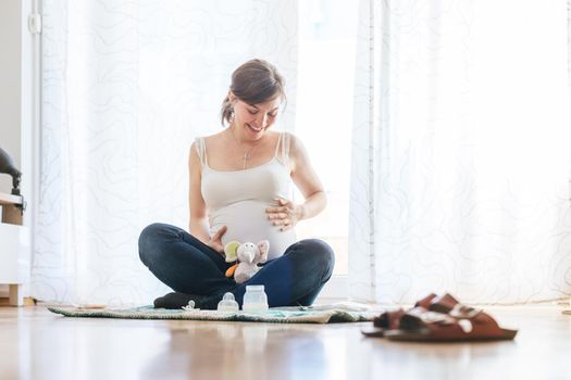 Happy Caucasian pregnant mother is sitting on the floor, touching her tummy, blue jeans