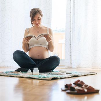 Happy Caucasian pregnant mother is sitting on the floor, reading about pregnancy, blue jeans