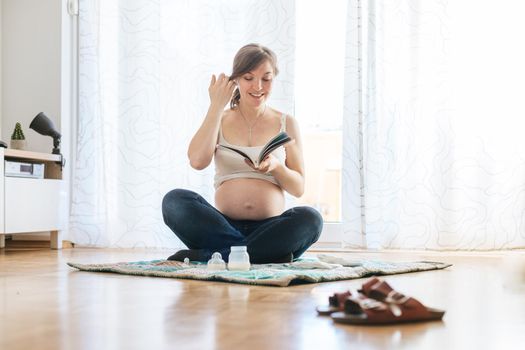 Happy Caucasian pregnant mother is sitting on the floor, reading about pregnancy, blue jeans