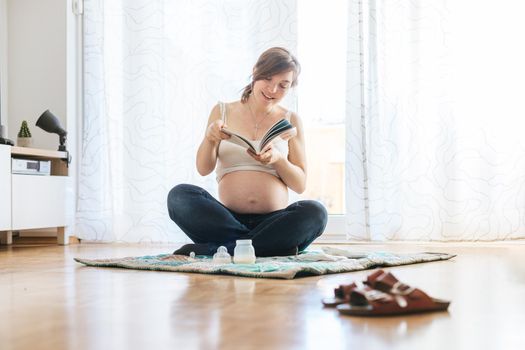 Happy Caucasian pregnant mother is sitting on the floor, reading about pregnancy, blue jeans