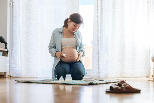 Happy Caucasian pregnant mother is sitting on the floor, touching her tummy, blue jeans