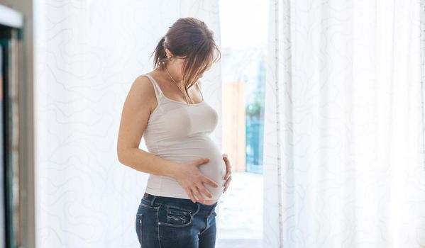 Happy Caucasian pregnant mother touching her tummy, blue jeans, close up