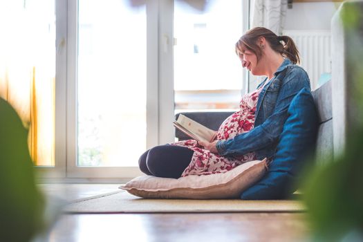 Pregnant caucasian happy mother is sitting on the floor, reading a book