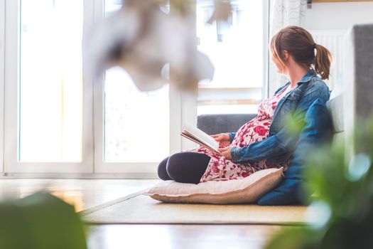 Pregnant caucasian happy mother is sitting on the floor, reading a book