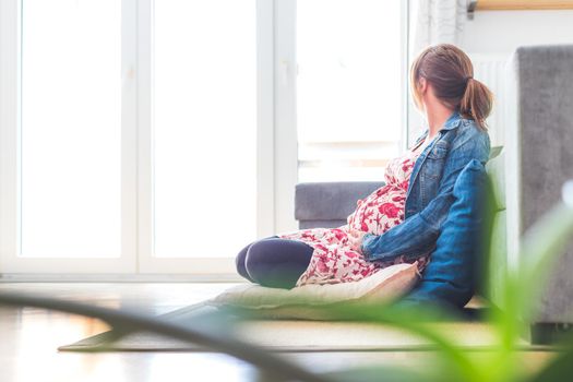 Pregnant caucasian happy mother is sitting on the floor, touching her tummy, floral dress