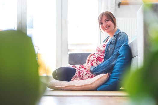 Pregnant caucasian happy mother is sitting on the floor, touching her tummy, floral dress
