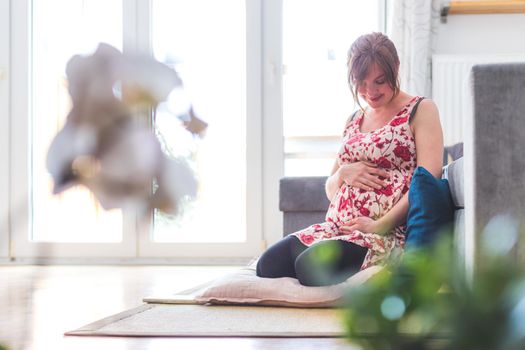 Pregnant caucasian happy mother is sitting on the floor, touching her tummy, floral dress