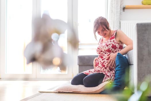 Pregnant caucasian happy mother is sitting on the floor, touching her tummy, floral dress