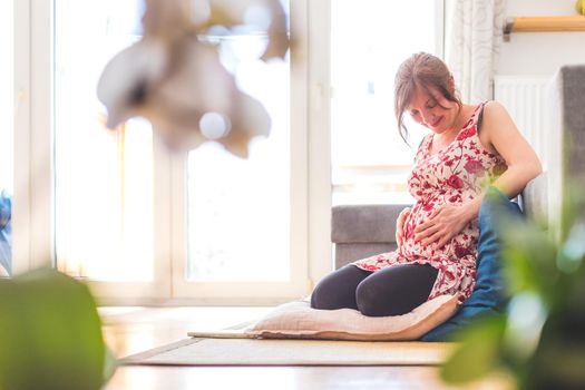 Pregnant caucasian happy mother is sitting on the floor, touching her tummy, floral dress