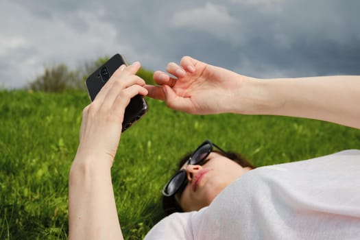 Girl with glasses lies on green grass and holds a smartphone.