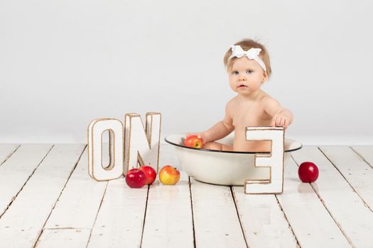 One-year-old child bathes in a basin. Beautiful baby girl with apples. First birthday girl.