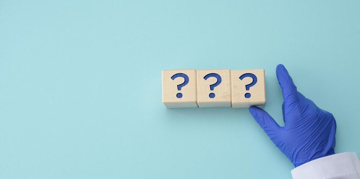 a hand in a blue medical glove holds cubes with question marks on a blue background. The concept of unknown, riddle, unanswered questions, copy space