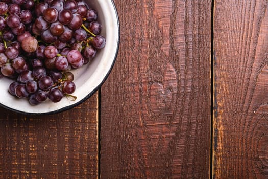 Fresh ripe grape berries in bowl on brown wooden background, top view copy space