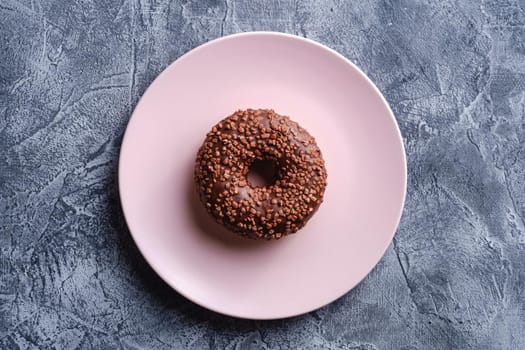 Chocolate donut with sprinkles on pink plate, sweet glazed dessert food on concrete textured background, top view