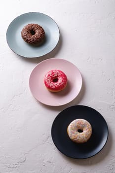 Three donuts in row on plates, chocolate, pink and vanilla donut with sprinkles, sweet glazed dessert food on white concrete textured background, angle view