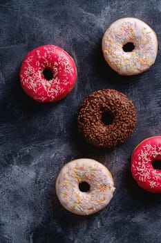 Chocolate, pink and vanilla donuts with sprinkles, sweet glazed dessert food on dark concrete textured background, top view