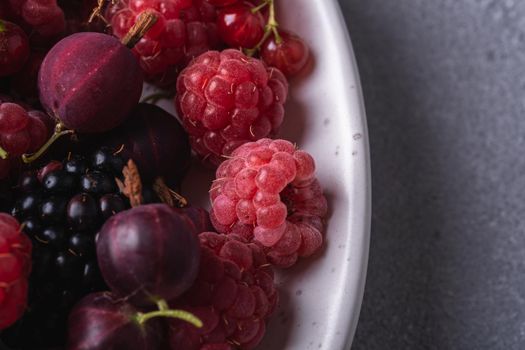 Tasty fresh ripe raspberry, blackberry, gooseberry and red currant berries in plate, healthy food fruit on stone concrete background, angle view macro