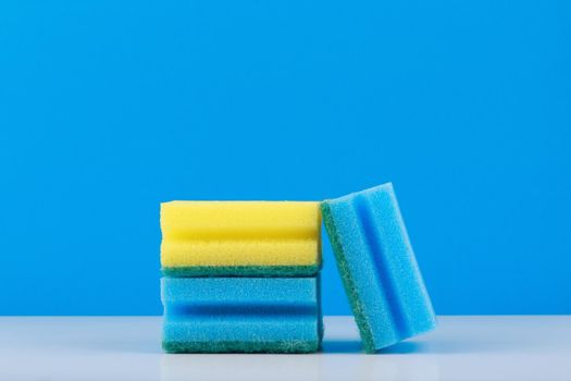 Blue and yellow sponge for cleaning and dish washing on white table against blue background with copy space. Concept of household equipment