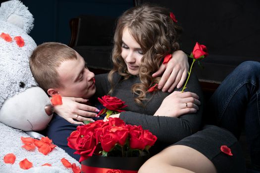 Beautiful young couple at home. Hugs, kisses and enjoys spending time together, celebrating Valentine's Day with red roses on a teddy bear.