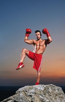 Vertical full length shot of a strong muscular male boxer posing outdoors on sunset wearing boxing gloves champion power strength fighter masculinity sports motivation competitive concept.