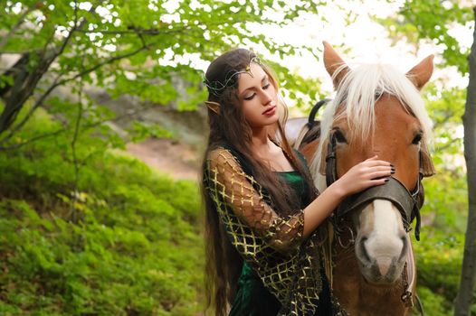 Beautiful young female elf with long dark wavy hair petting her horse resting in the woods forest nymph stroking her horse care pet love animals harmony caring owner gentle creature myth legend movie.