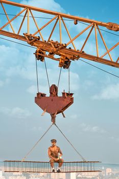 View from distance of bold builder in work wear sitting on construction on high and eating. Building crane holding construction with man over city. Extreme building of house in big city.