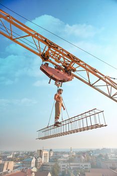 Side view of athletic man in hat standing on construction on high and looking away. Large building crane holding construction with male over city in air. Cityscape and blue sky on background.