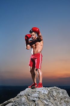 Handsome young male fighter with stunning hot muscular ripped body wearing boxing gloves training outdoors on sunset determination practicing workout exercising preparing victory fighting concept