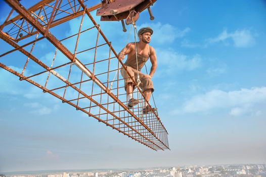 Iron building crane holding construction with athletic man in work wear on high. Male standing on construction and looking at distance away. Extreme building in big city.