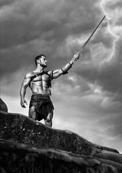 No one he fears. Monochrome low angle shot of a fearless muscular warrior with a sword in his hand posing under stormy sky on a mountain peak