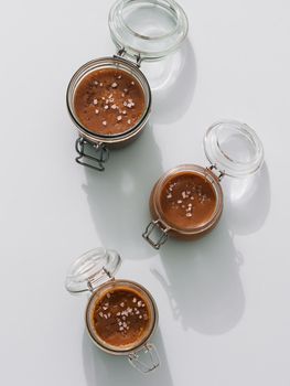 Salted caramel in glass jars, top view. Brown caramel or condensed milk with sea salt crystalls, shoot in bright or hard light. Light neutral background. Vertical