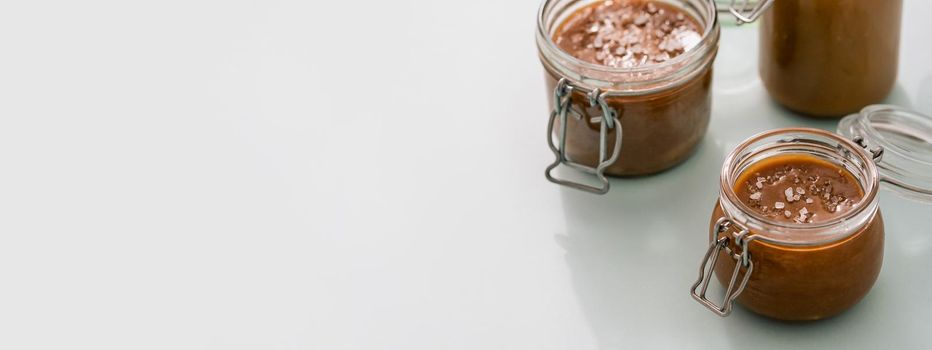 Salted caramel in glass jars on light neutral background. Brown caramel or condensed milk with sea salt crystalls. Long horizontal banner, copy space left