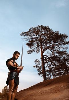 Getting thoughts together. Low angle vertical portrait of a young handsome gladiator warrior praying with a sword in his hands