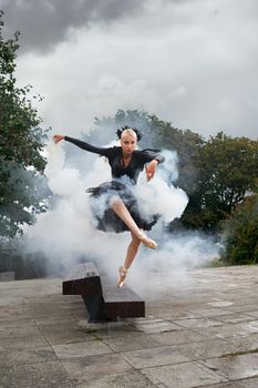 Expressive performance of a black swan ballerina. Beautiful blonde female ballet dancer jumping in the air while dancing in the smoke outdoors.