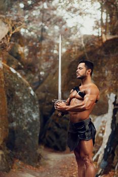 Soul of a fighter. Vertical portrait of a handsome young gladiator with stunning muscular torso posing with a sword near the rocks in the forest