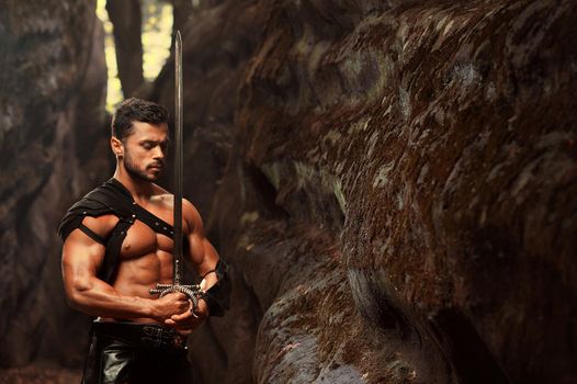 Deep in his thoughts. Horizontal portrait of well-built muscular young warrior standing with a sword with his eyes closed near the rock at the woods