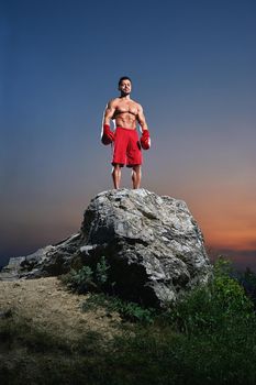 Young muscular male fighter wearing boxing gloves standing in a stance on a rock outdoors on sunset copyspace people lifestyle sports athletics sportsman fitness muscles combat martial fighting.