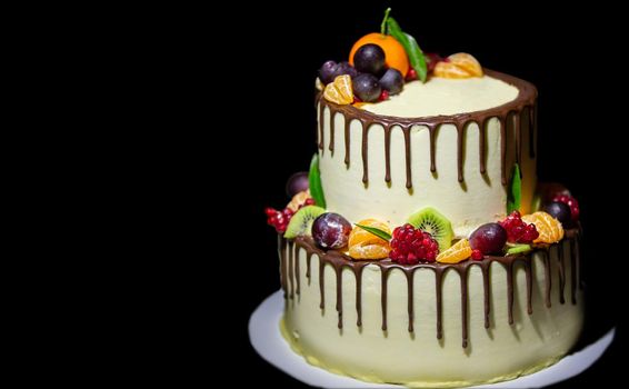Beautiful two-tier cake poured with chocolate and decorated with fresh fruits on a black background.