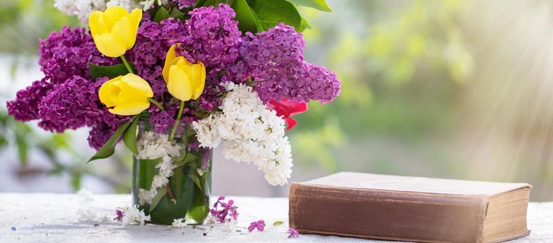 Spring background. Lilac and tulips in a glass jar on the table, also an old book. Beautiful flowers lit by the rays of the sun.