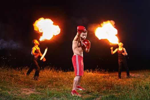 Full length shot of a professional boxer posing in fighting stance outdoors at night exotic fire show on the background fire eaters blowing fire from their mouths epic show time champion victory sport.
