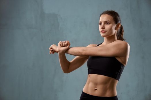 Front view of fit caucasian female model stretching arm, wearing black sportswear standing and looking aside, isolated on loft gray studio background. Muscular pretty young fitnesswoman.